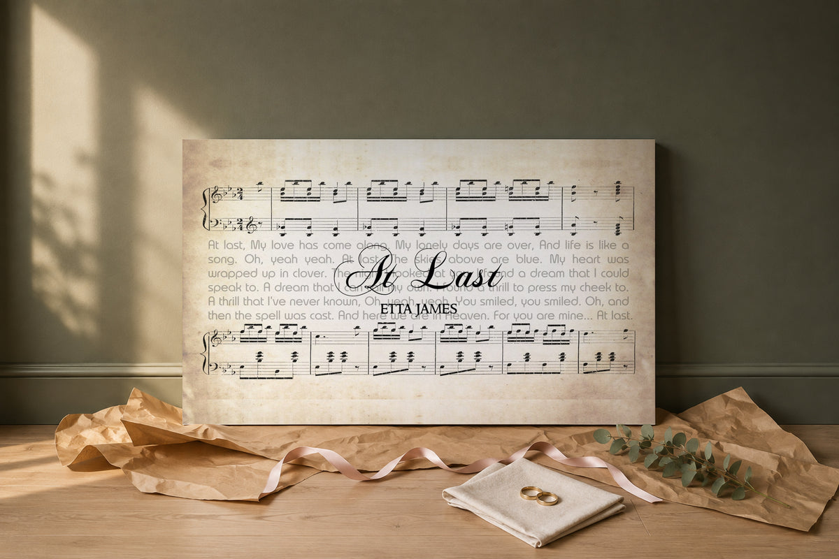 At Last lyric canvas as a wedding gift, unwrapped on a pale oak floor with kraft paper, blush ribbon, gold wedding bands and eucalyptus