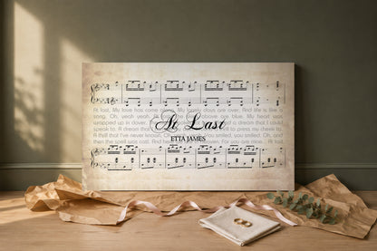At Last lyric canvas as a wedding gift, unwrapped on a pale oak floor with kraft paper, blush ribbon, gold wedding bands and eucalyptus
