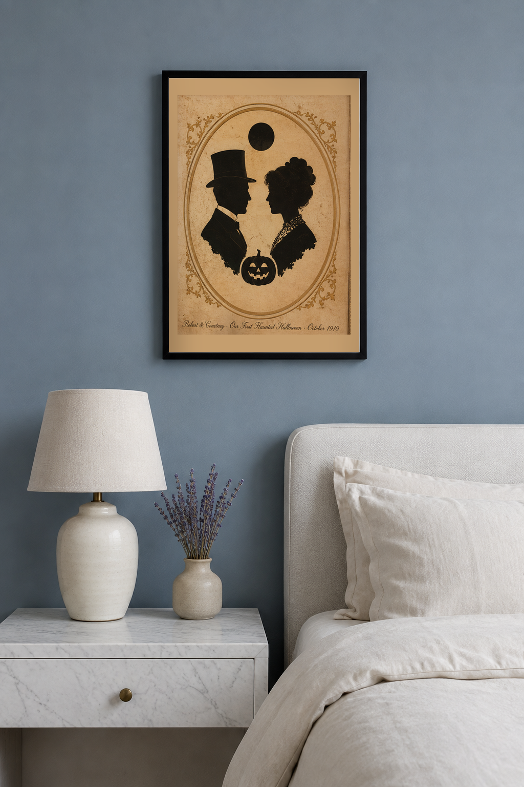 Personalized couple names and date Halloween print in a black frame on a dusty blue grey bedroom wall above a white marble nightstand with a linen lamp and dried lavender