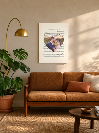 Custom song lyrics sheet music print with wedding photo heart, hung in a cozy mid-century living room above a caramel sofa with a brass floor lamp and monstera