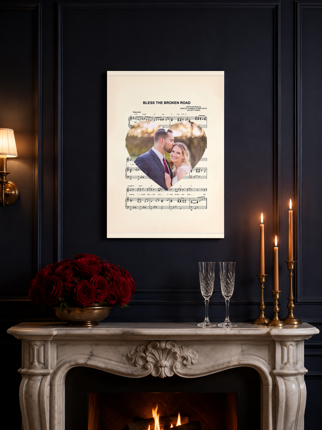 Personalized first dance sheet music print displayed above a white marble anniversary mantel with red roses, champagne flutes and brass taper candles