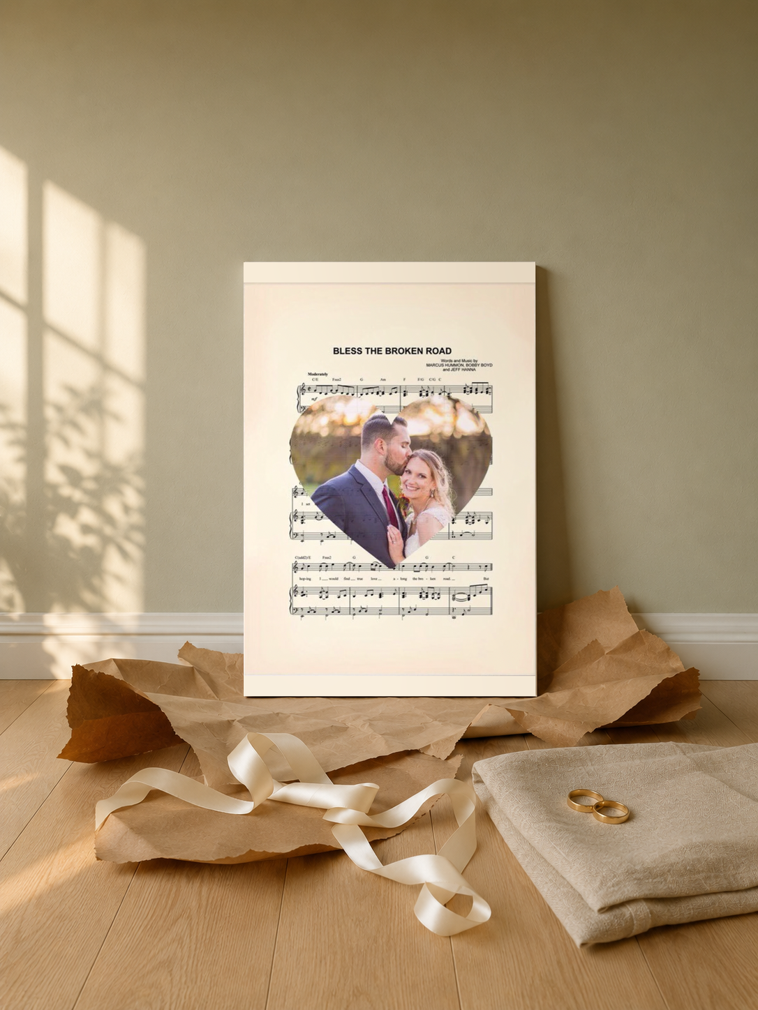 Custom first dance sheet music print unwrapped as a wedding gift, leaning on an oak floor with kraft paper, satin ribbon and gold wedding bands